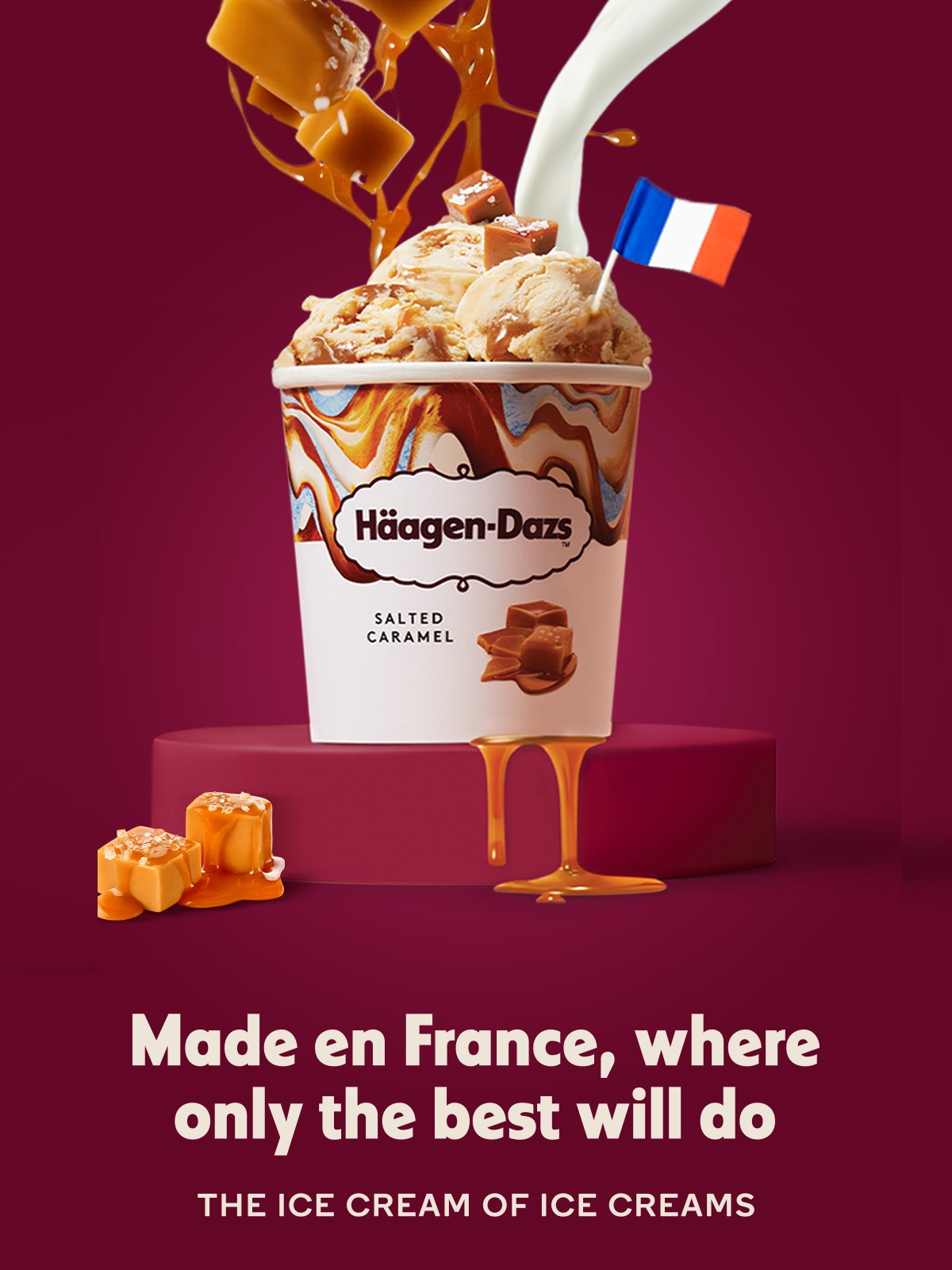 A luxurious advertisement for Häagen-Dazs Salted Caramel ice cream featuring a cup overflowing with scoops of ice cream, rich caramel sauce, and caramel pieces. The caramel sauce drips down the sides onto a pedestal, enhancing the indulgent appeal. A small French flag is placed on top, highlighting the product’s French origin. The background is a deep burgundy, with the tagline “Made en France, where only the best will do” and “THE ICE CREAM OF ICE CREAMS” displayed below.